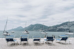 Ein Kiesstrand mit Reihen blauer und weißer Liegestühle, vor einem ruhigen See mit Segelbooten und Bergen im Hintergrund.