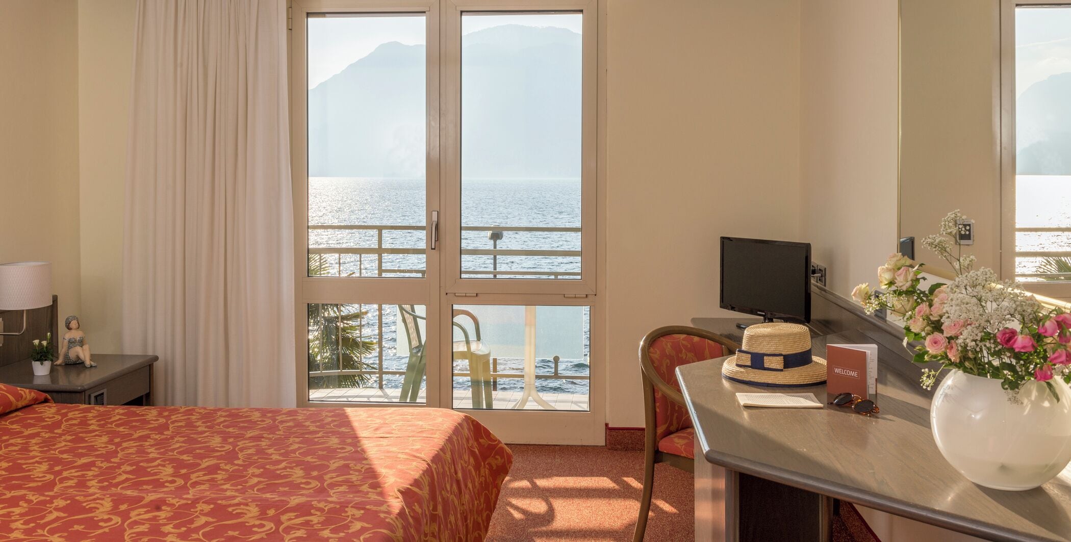 A hotel room with a bed, a desk with a computer, a chair, a window with a view of the sea, and a vase with flowers.
