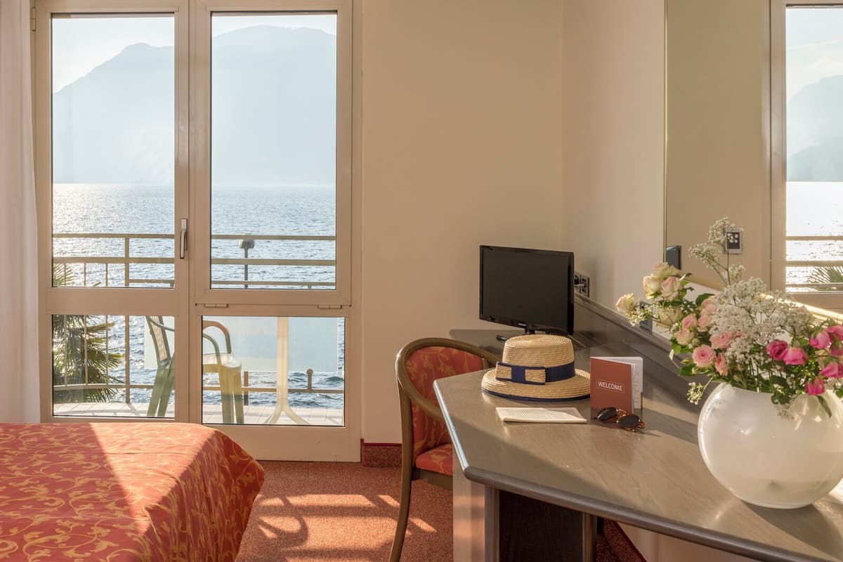 Ein Hotelzimmer mit einem Bett, einem Schreibtisch mit Computer, einem Sessel, einem Fenster mit Meerblick und einer Vase mit Blumen.