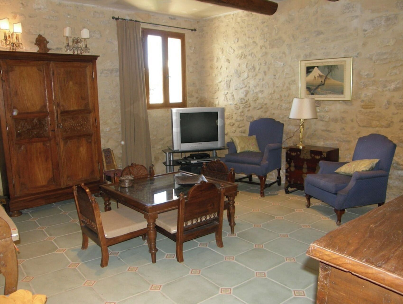 A living room with a stone wall, a wooden cabinet, a television, two blue armchairs, and a dining table with chairs.
