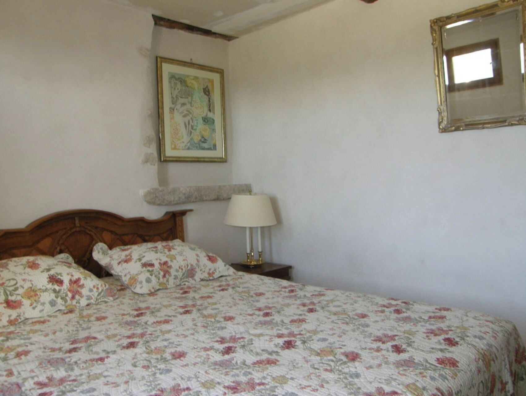 Una camera da letto con un letto di legno, biancheria da letto floreale, una lampada da comodino, un quadro appeso al muro e uno specchio.