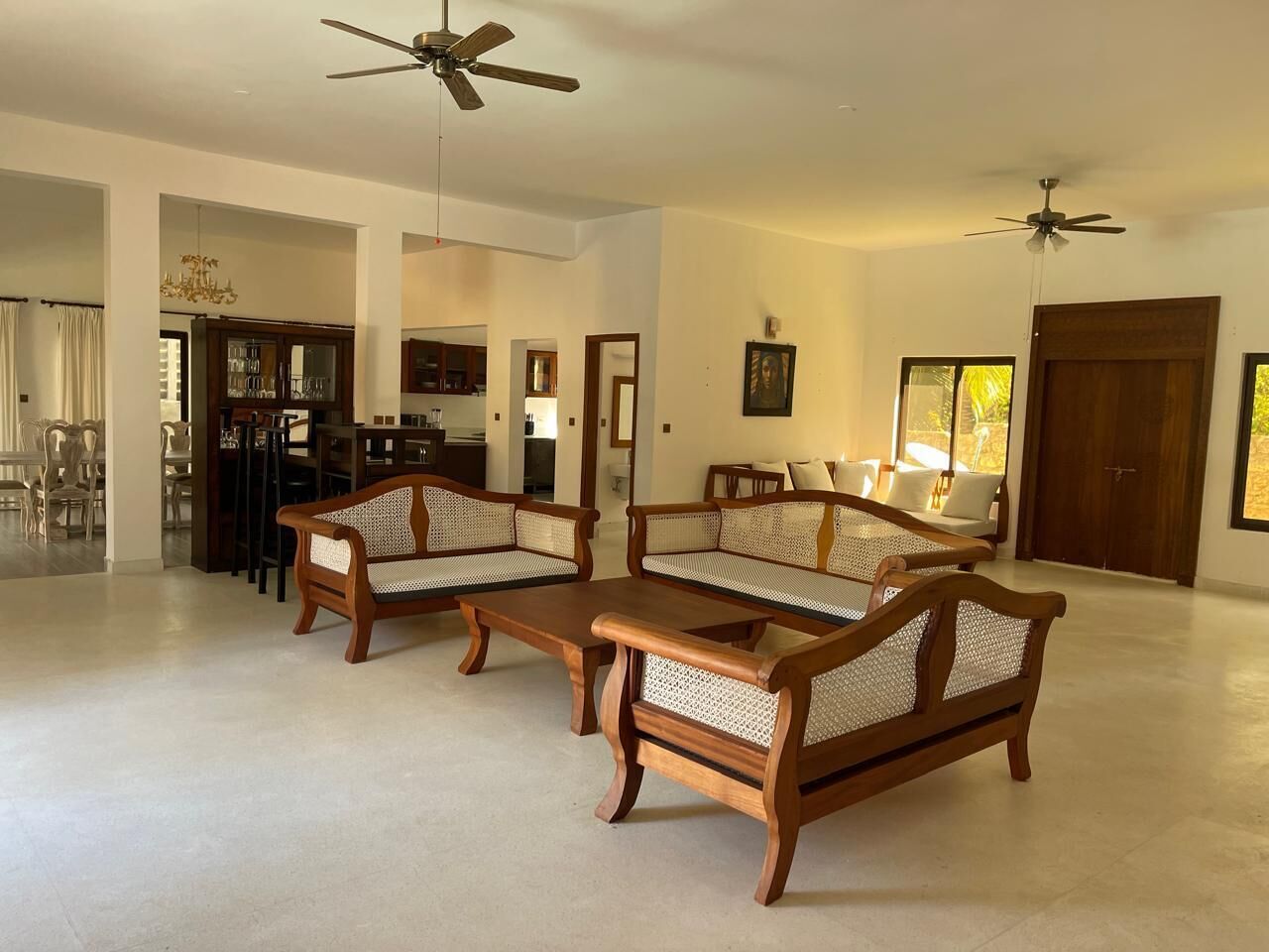A spacious living room with two wooden sofas and a coffee table, a ceiling fan, and a door leading to another room.
