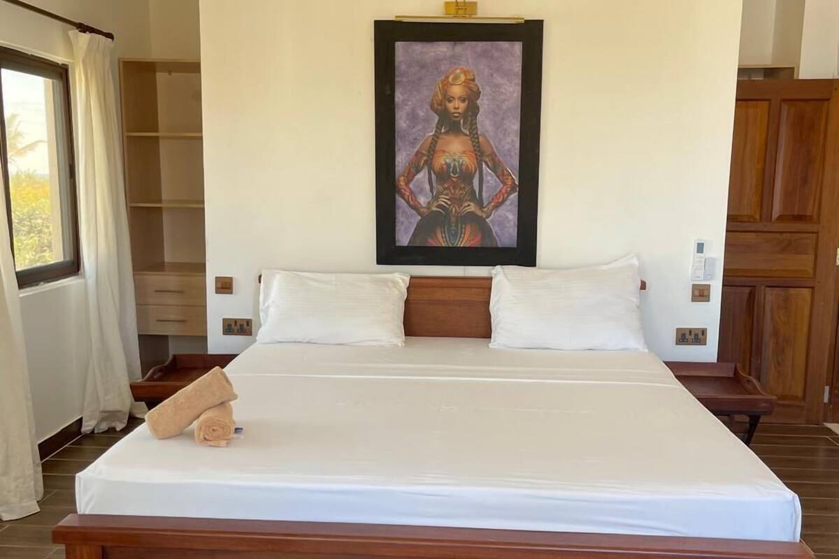 Uma cama grande com estrutura de madeira, roupa de cama branca e uma toalha enrolada no rodapé. Uma obra de arte emoldurada está centralizada acima da cama.