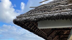 Un bâtiment moderne doté d’un balcon en verre donnant sur une étendue d’eau, d’un toit de chaume et offrant une vue dégagée sur la terre ferme au loin.