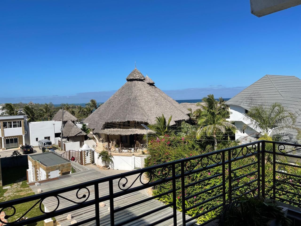 A thatched-roof structure with a modern building in the background, surrounded by greenery and a clear sky.