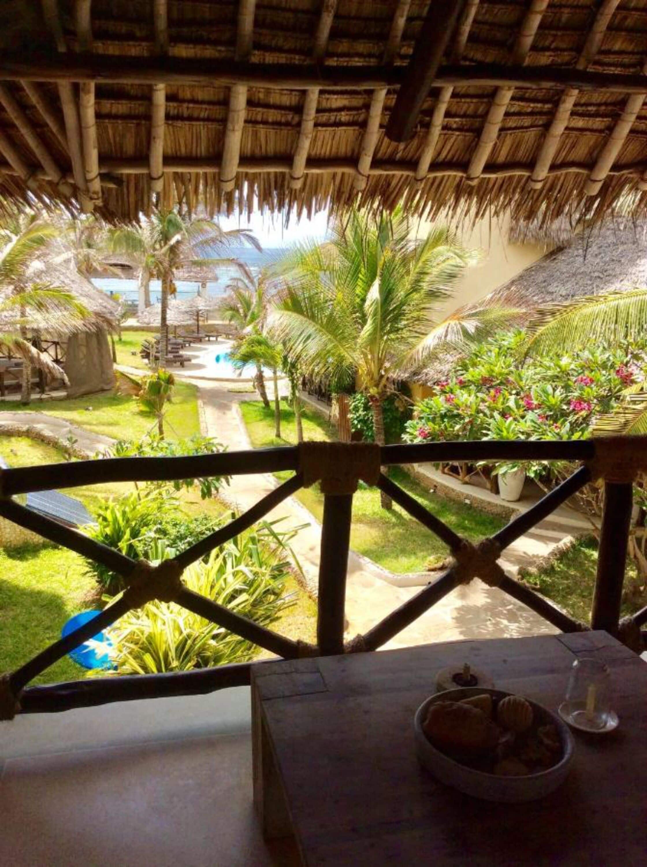 Un espacio al aire libre con techo de paja, piscina, palmeras y una mesa de comedor con un tazón y una copa.