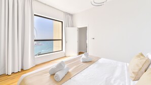 Een moderne hotelkamer met een groot raam met uitzicht op de zee en een reuzenrad, een net opgemaakt bed met witte lakens en een houten vloer.