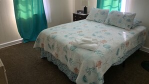 A neatly made bed with floral bedding, a bedside table with a lamp, and curtains. - Queen Anne's Revenge At The Beach 3 Bedroom Cottage by RedAwning (Kitty Hawk)