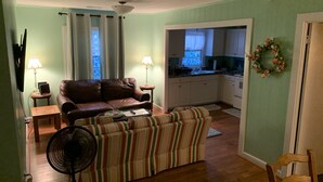 A living room with a brown leather sofa, a striped armchair, and a fan. There is a kitchen visible through an open doorway. - Queen Anne's Revenge At The Beach 3 Bedroom Cottage by RedAwning (Kitty Hawk)