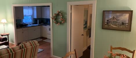 A living room with a striped sofa, a wooden dining table with a vase of flowers, a kitchen in the background, and a door with a wreath.