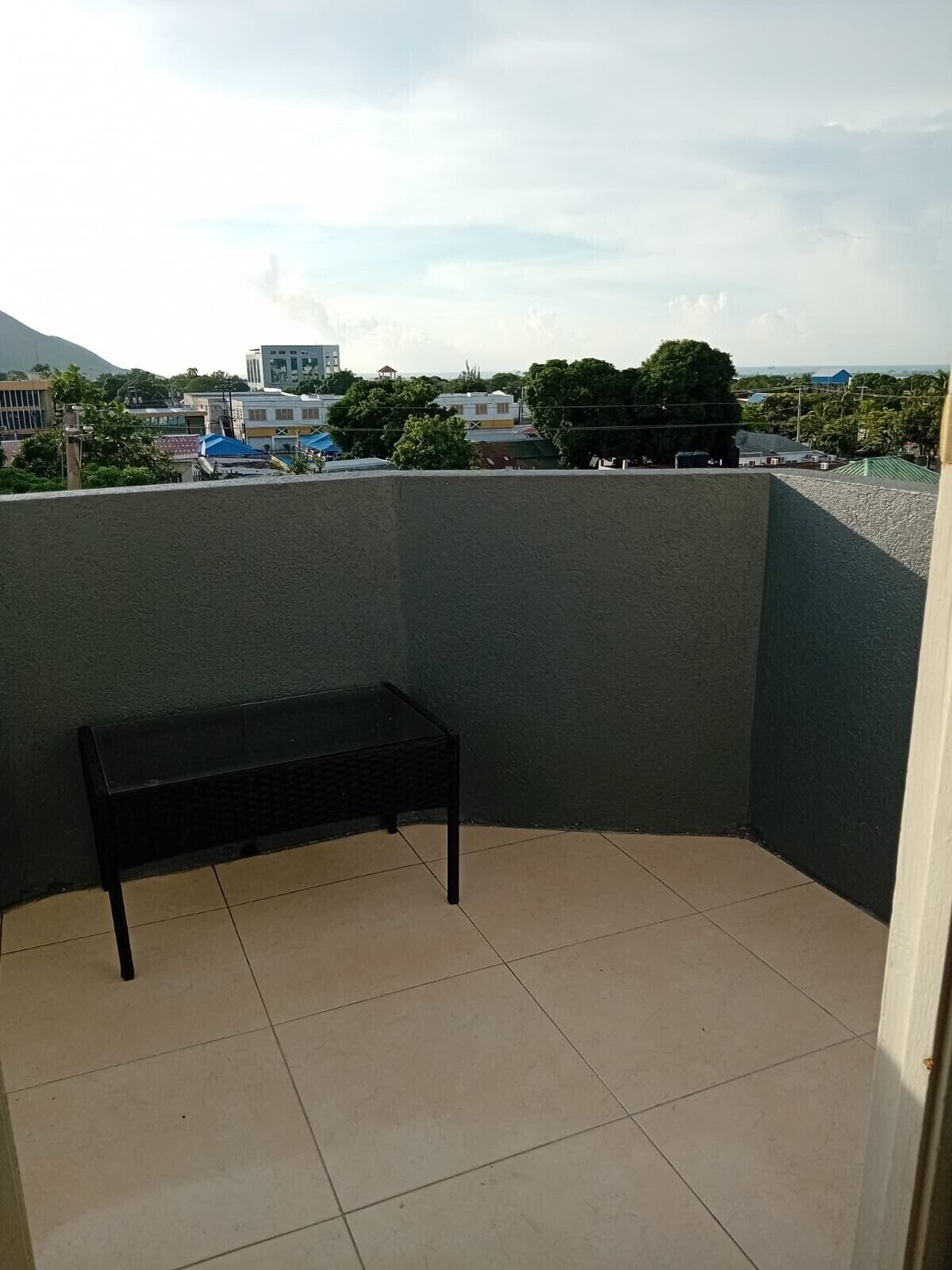 A corner balcony with a black bench, tiled floor, and a view of buildings and trees in the background.