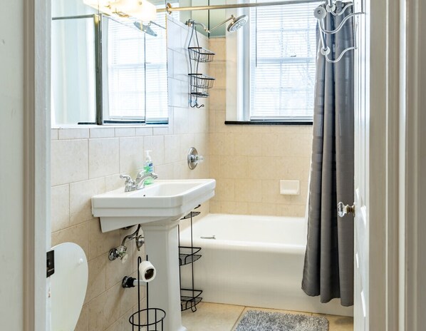 A bathroom with a white sink, a bathtub, and a shower. There are windows with blinds, a towel rack, and a shower curtain.