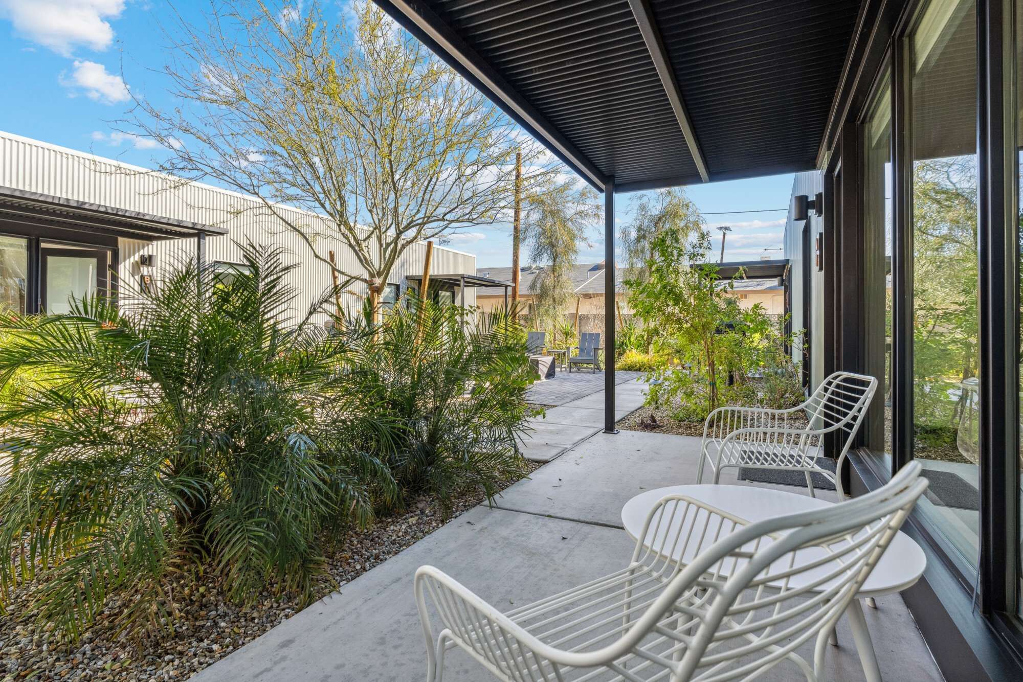 A covered patio with outdoor seating, surrounded by greenery and a view of neighboring buildings.