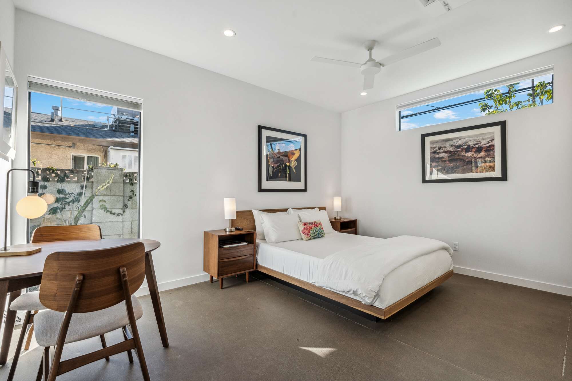 A modern bedroom with a bed, bedside tables, a desk, and chairs. There are framed pictures on the walls and a view of the outside through a window.