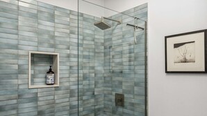 Une salle de bain avec un mur de douche recouvert de carreaux de verre, un tableau encadré au mur et un distributeur de savon intégré dans un renfoncement.