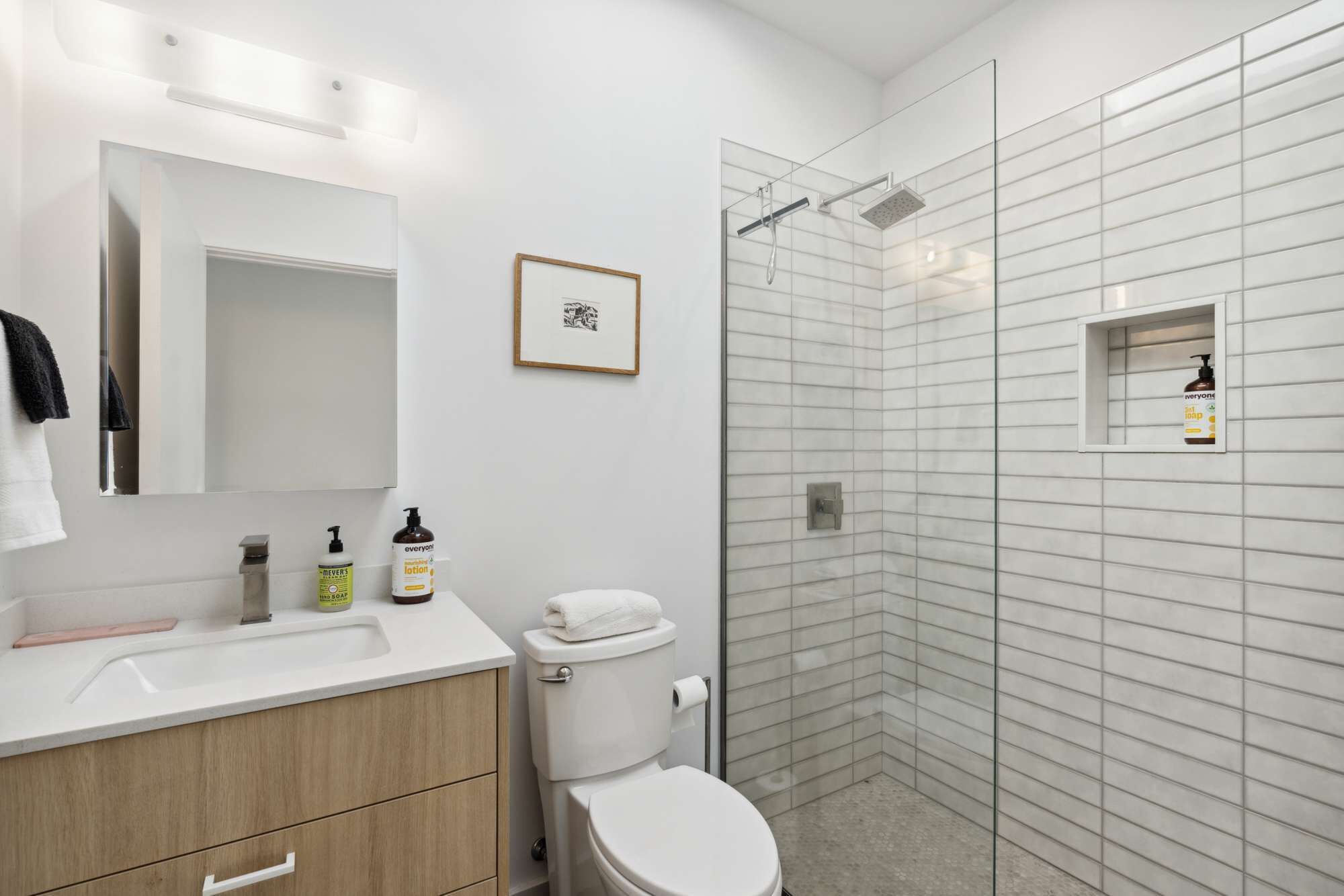 Ein modernes Badezimmer mit Waschbecken, Spiegel und Toilette in Weiß. An der Wand hängt ein gerahmtes Bild und es gibt eine Dusche mit Glastür.