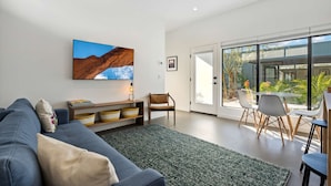 A modern living room with a large flat-screen TV, a sofa, a coffee table, and a dining area with chairs and a table. - Unique Design 1-bedroom Container - 80 Walkscore! (Phoenix)