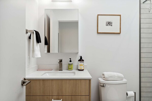 Une salle de bain moderne avec un lavabo blanc, un miroir et des toilettes. On trouve des articles de toilette sur le lavabo et un tableau encadré au mur.