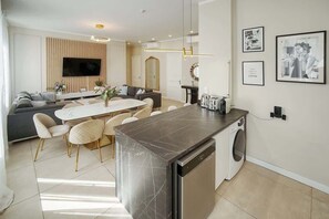 A modern kitchen with a built-in dishwasher, a dining area with a marble table, and a living room with a sofa and TV.