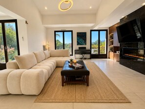 A modern living room with a sectional sofa, a flat-screen TV, a fireplace, and a large window offering a view of greenery.