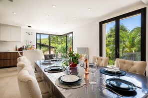 Un espace repas moderne avec une longue table dressée pour six personnes, de grandes fenêtres donnant sur un paysage verdoyant et une cuisine dotée de meubles blancs.