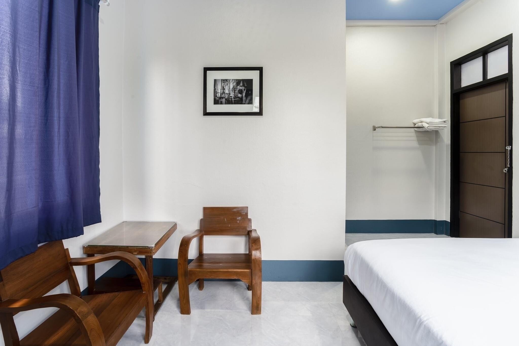A hotel room with a bed, a small table, two wooden chairs, a framed picture on the wall, and a towel rack.