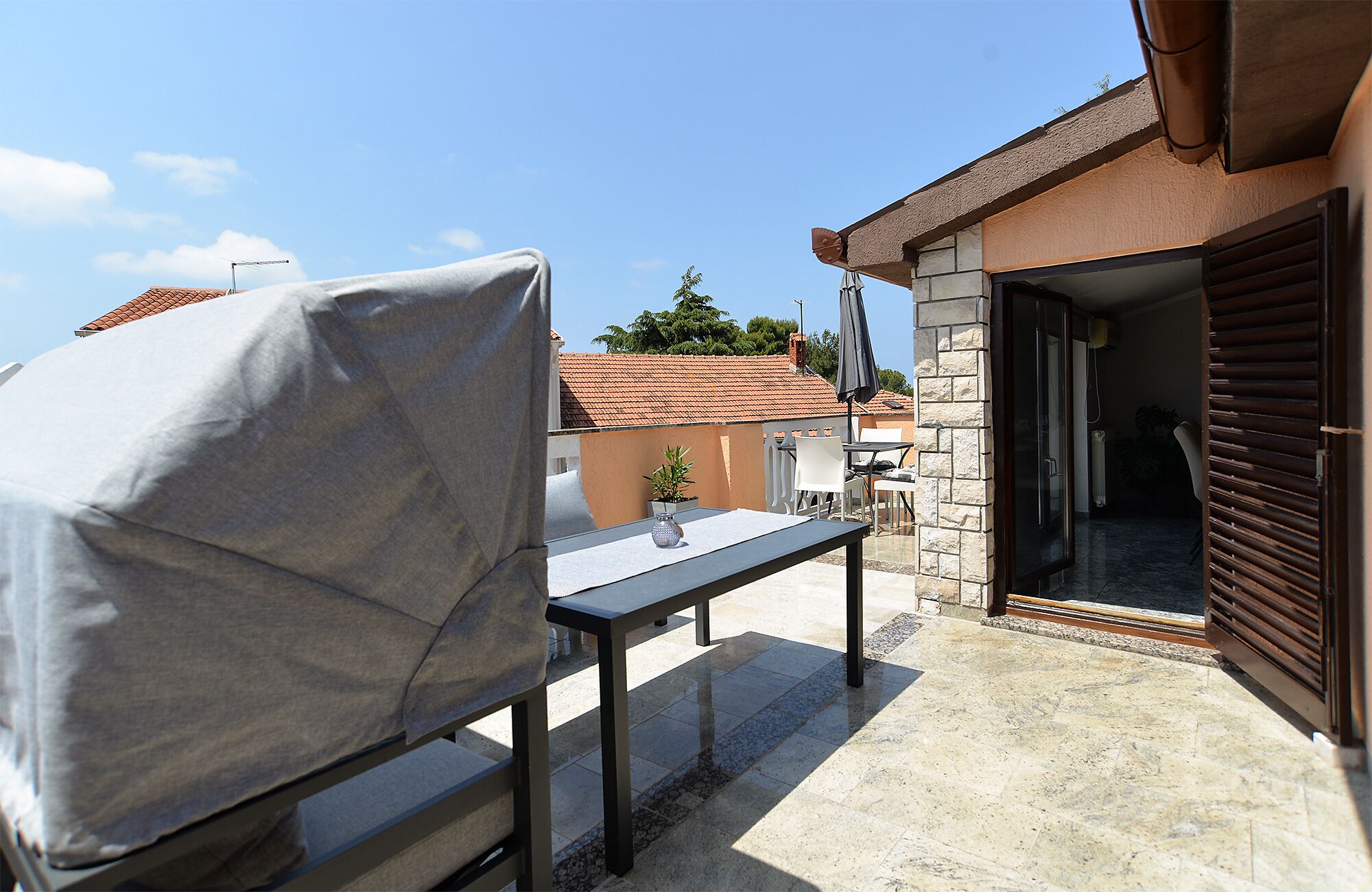 An outdoor patio with a covered table and chairs, a stone wall, and a view of neighboring buildings.