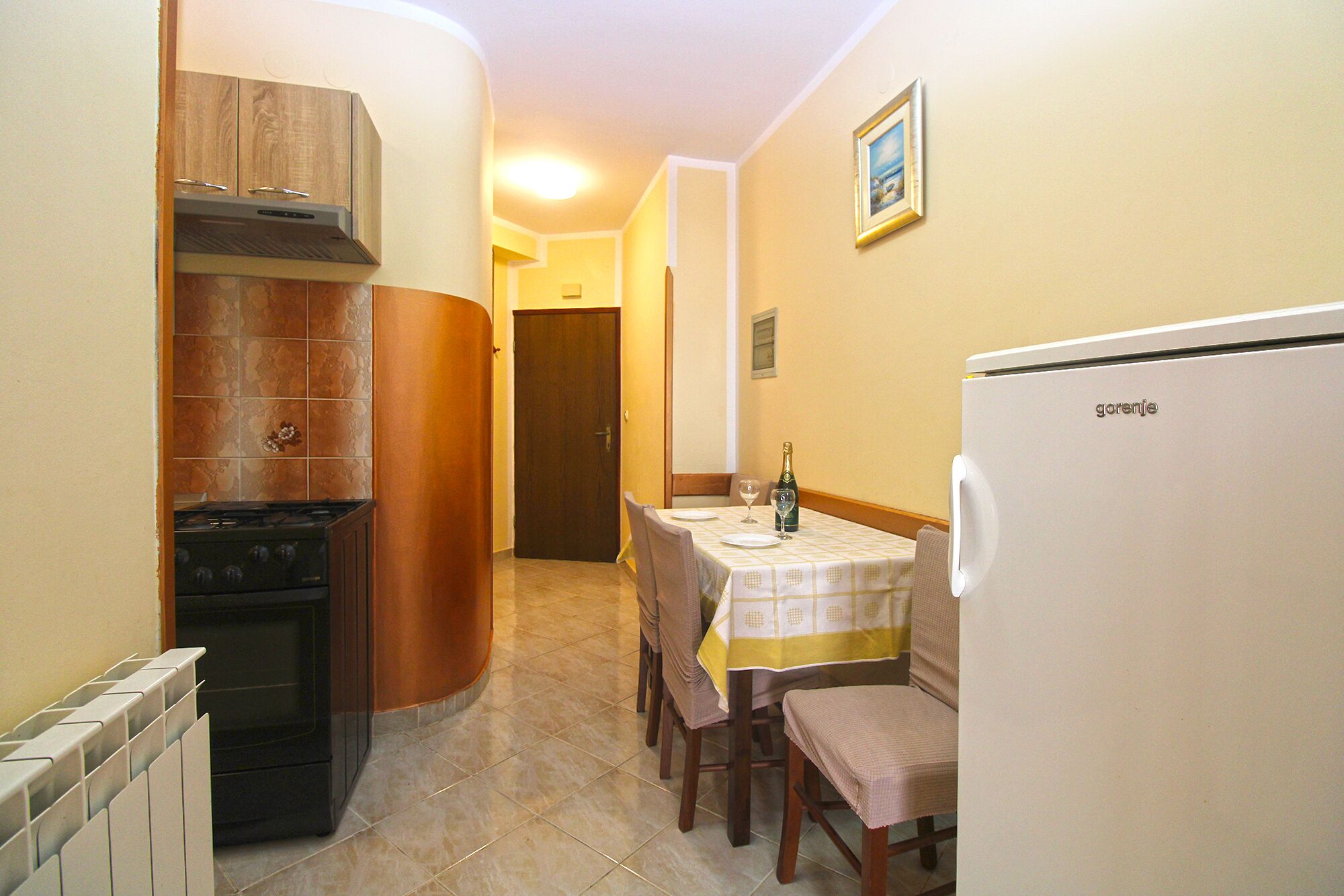 A compact kitchen with a stove, oven, and a refrigerator. There is a dining table with chairs and a bottle of champagne.