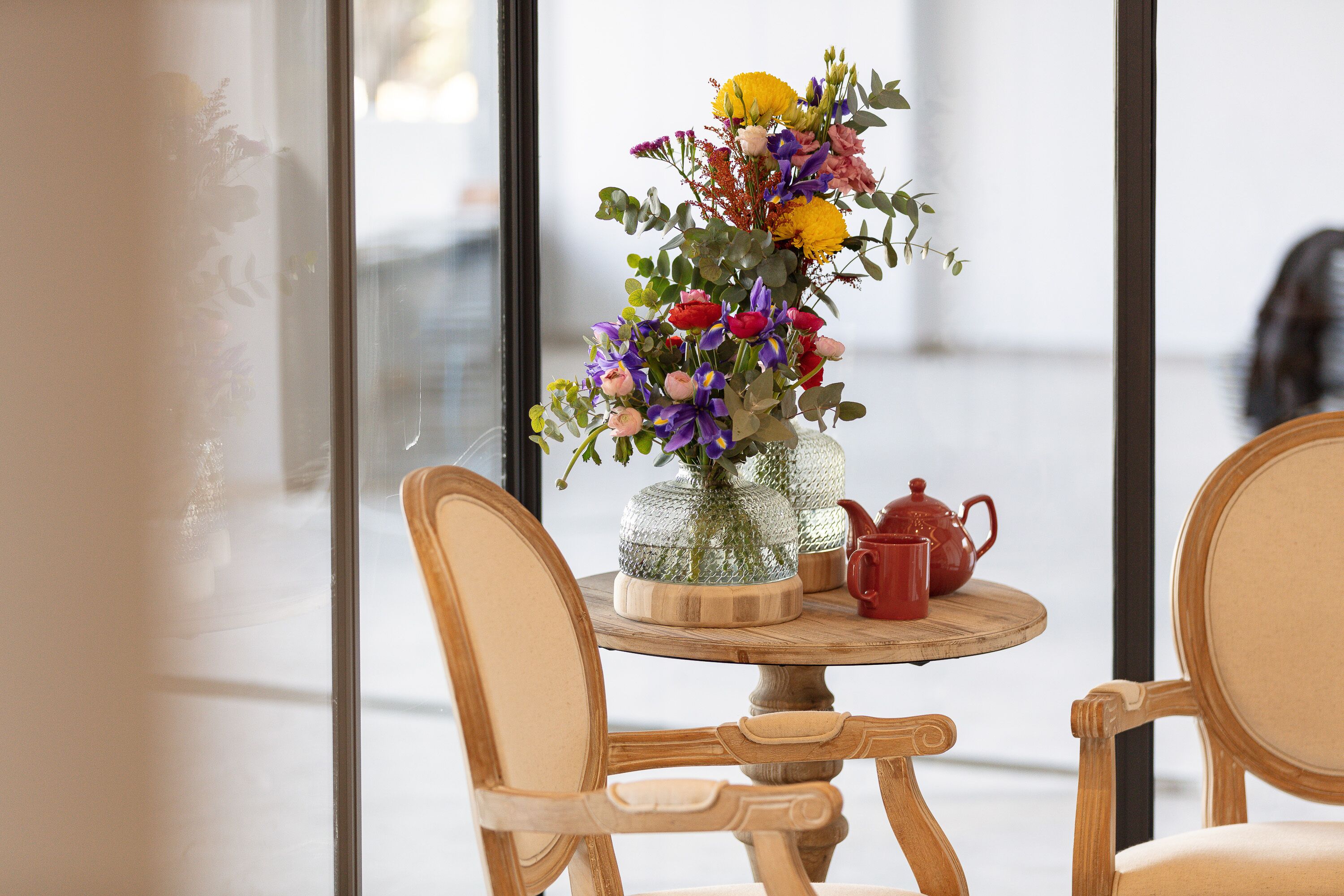 Una mesa redonda de madera con un jarrón de flores, una tetera roja y un recipiente de vidrio. Dos sillas de madera están colocadas alrededor de la mesa.