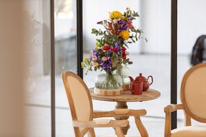 Uma mesa redonda de madeira com um vaso de flores, uma chaleira vermelha e um recipiente de vidro. Duas cadeiras de madeira estão dispostas ao redor da mesa.