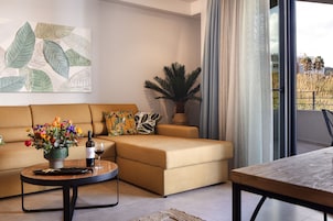 A modern living room with a beige sofa, a coffee table with a vase of flowers, a wine bottle, and a glass. There is a large window with curtains, a potted plant, and a view of buildings outside.