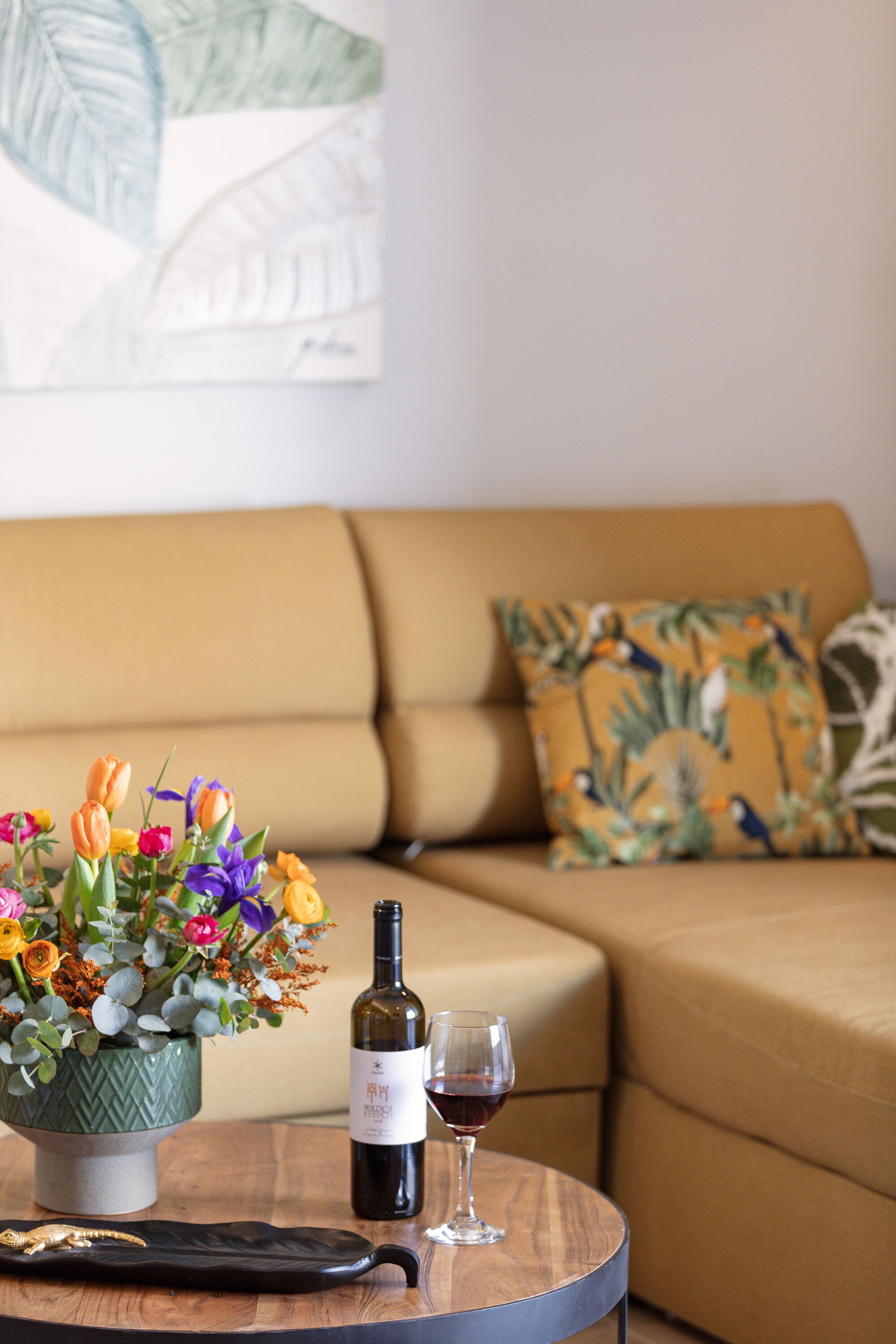 Een ronde houten tafel met een fles wijn en een glas, een bloemenvaas vol kleurrijke bloemen en een decoratief bord.