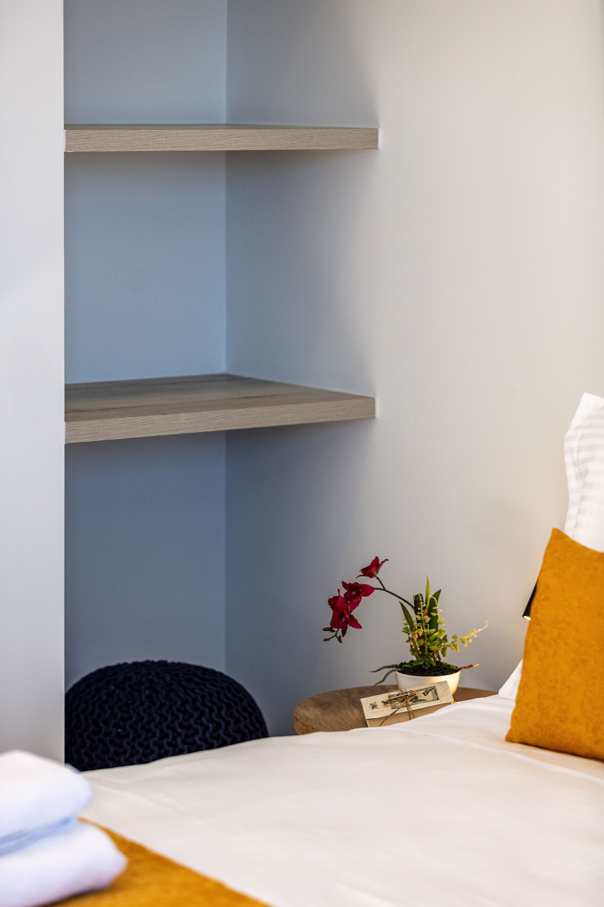 A hotel room with a bed, a nightstand, a potted plant, and a wall-mounted shelf.