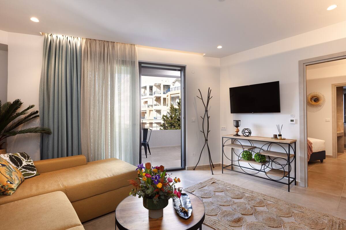 A modern living room with a sofa, a coffee table with a vase of flowers, a television mounted on the wall, and a view of the outside balcony.