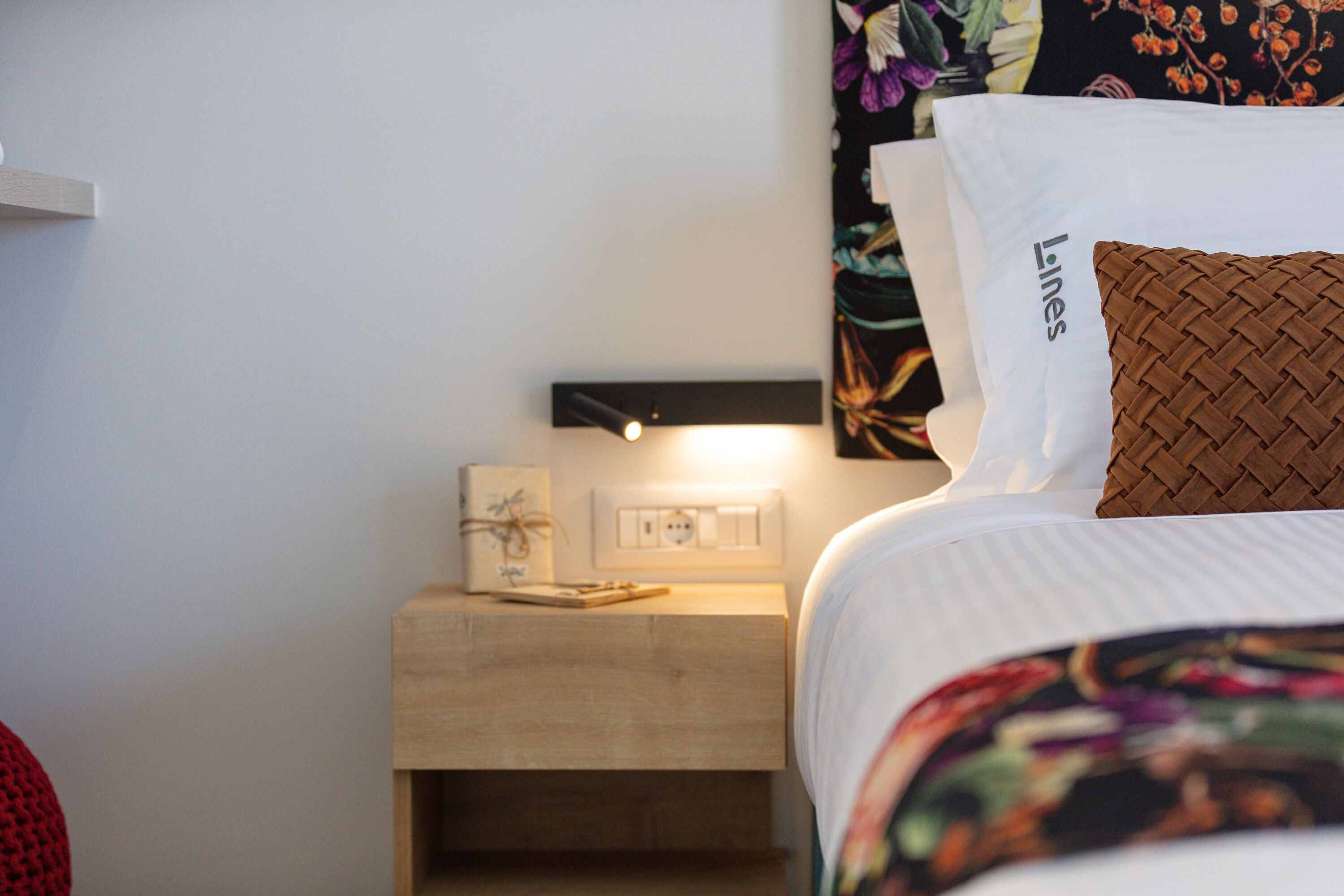 A neatly made bed with a pillow labeled "Lines" and a bedside table with a lamp and a framed picture.