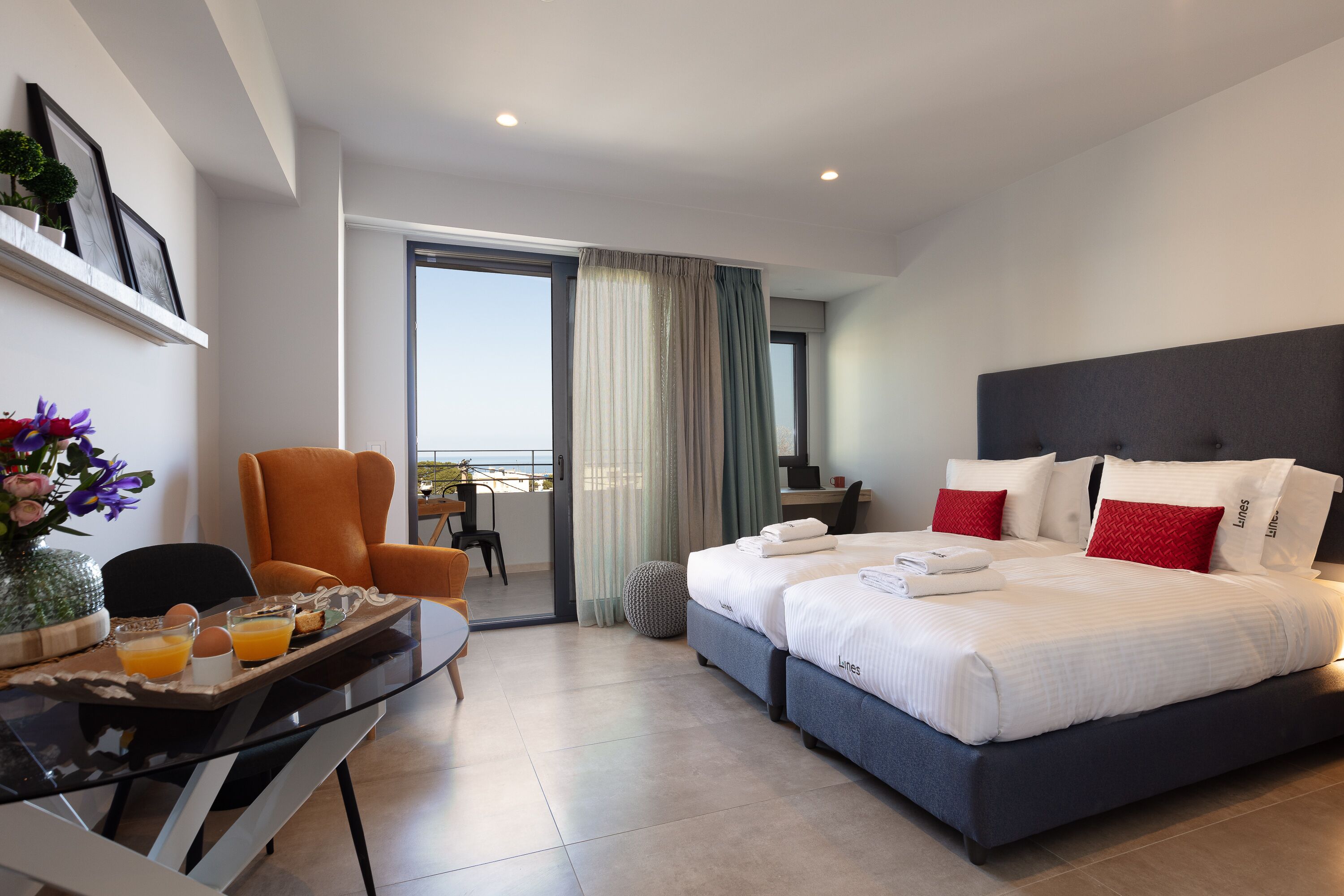 A modern hotel room with a large bed, a dining area with a tray of breakfast, a chair, and a view of the outdoors.