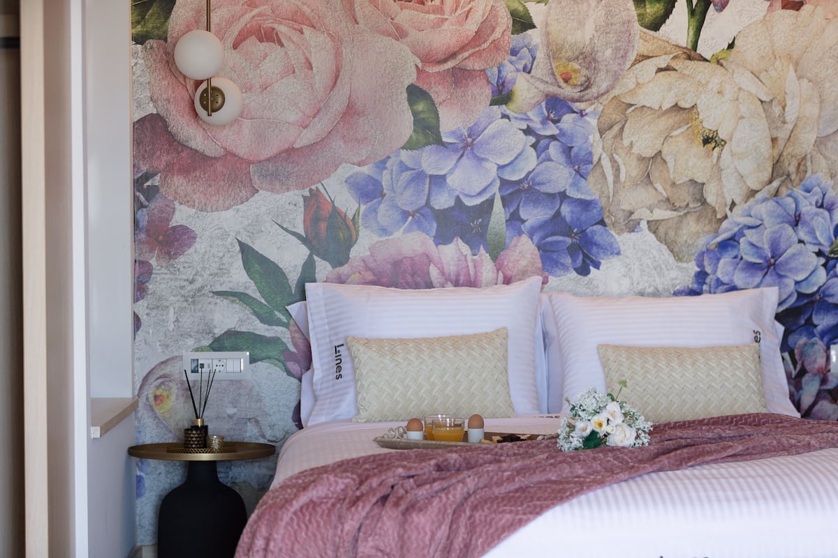 A bedroom with a floral wallpaper, two beds with pillows, a bedside table with a lamp, and a small table with a tray of eggs and juice.