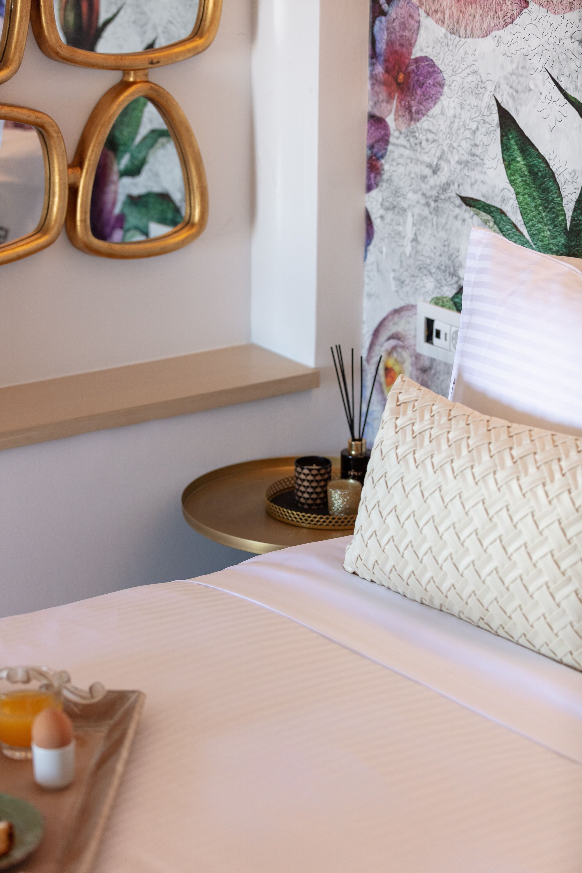 Una cama bien hecha con un almohadón blanco y uno tejido, una bandeja con un vaso de jugo de naranja y un huevo duro, y una pared con papel tapiz floral y un espejo.