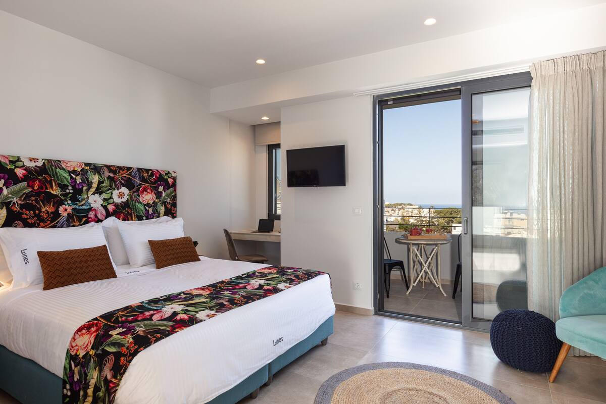 A modern hotel room with a large bed, a desk, a TV, and a balcony with a table and chairs.