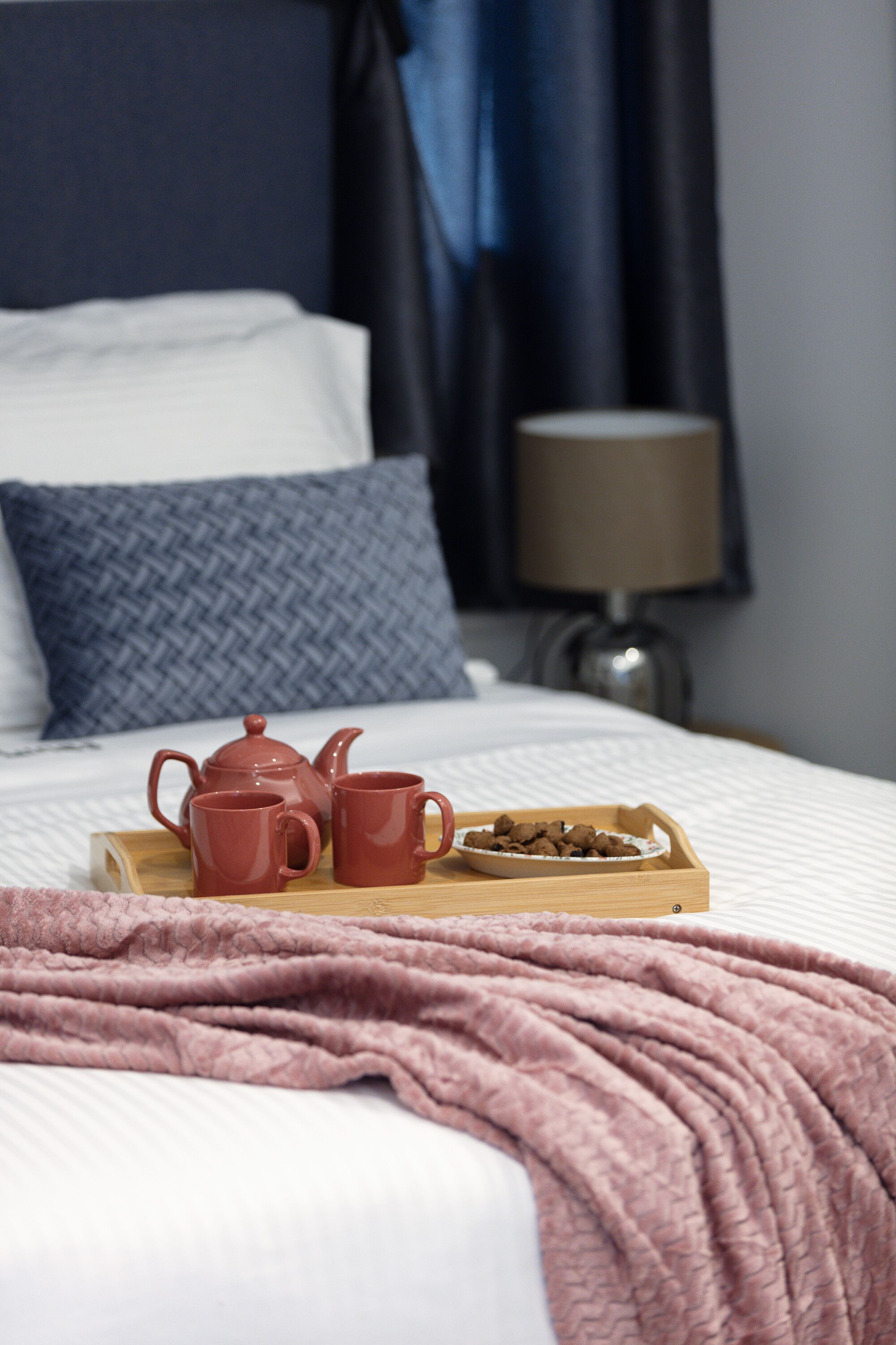 Un lit bien fait, recouvert d’une couverture rose, une tablette en bois avec une théière, deux tasses et un bol de céréales.