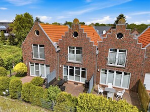 A two-story brick house with a sloped roof, multiple windows, and a patio area with outdoor furniture. - Schönes Reihenmittelhaus in Strandnähe (Norddeich)