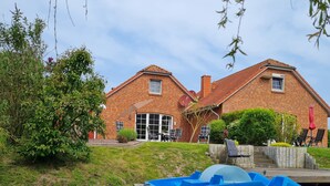 A house with a brick exterior, a large glass door, and a patio with chairs and an umbrella. - Wunderschönes Ferienhaus mit Eigenem Tretboot (Greetsiel)