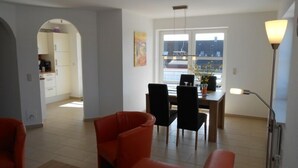 Ein modernes Wohnzimmer mit Essbereich, einer Küche im Hintergrund und einem großen Fenster, das natürliches Licht hereinlässt.