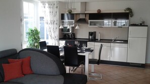 Uma sala de estar moderna com um sofá cinza, mesa de jantar e cadeiras. Uma cozinha com armários brancos e eletrodomésticos pretos é visível ao fundo.