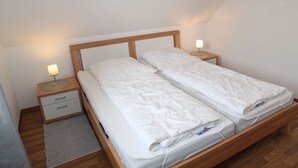 Uma cama de casal com cabeceira de madeira, duas mesas de cabeceira com abajures e um tapete cinza sobre piso de madeira.