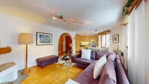 Uma sala de estar moderna com um sofá roxo, uma mesa de centro de vidro e piso de madeira. Cozinha visível ao fundo, com armários de madeira e uma janela.