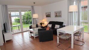 A modern living room with a black sofa, a white coffee table, and a dining table with chairs. There are two windows with curtains, and a TV on a stand. - Lichtdurchflutete Wohnung im Löwenpark (Norden)