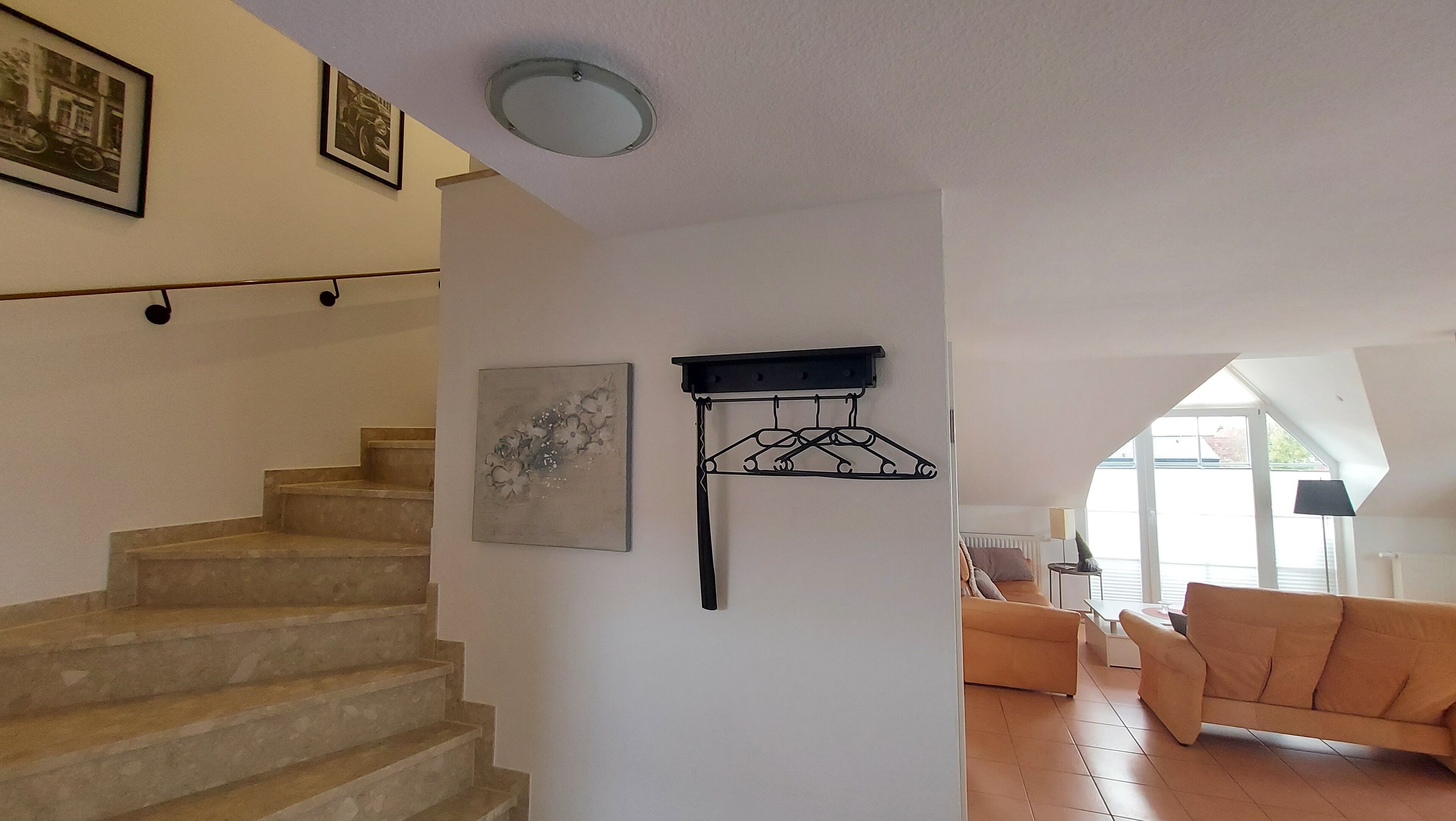 Una escala que porta a un segon pis, una prestatgeria de metall negre, una obra d'art emmarcada, un sofà i una làmpada en una habitació lluminosa amb grans finestres.
