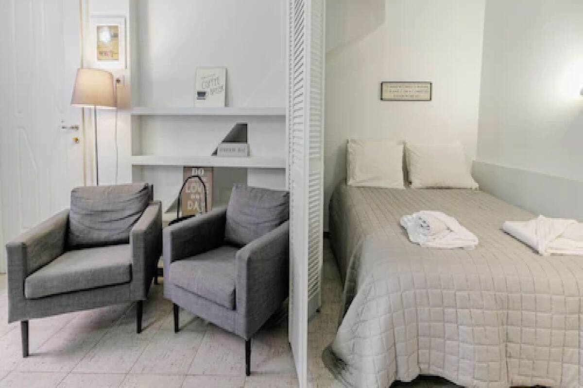 Une chambre à coucher comprenant un lit, deux fauteuils, une table de chevet, une lampe et une étagère avec des objets décoratifs.