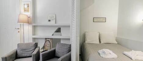 Un dormitorio con una cama, dos sillones, una mesita de noche, una lámpara y una estantería decorada.